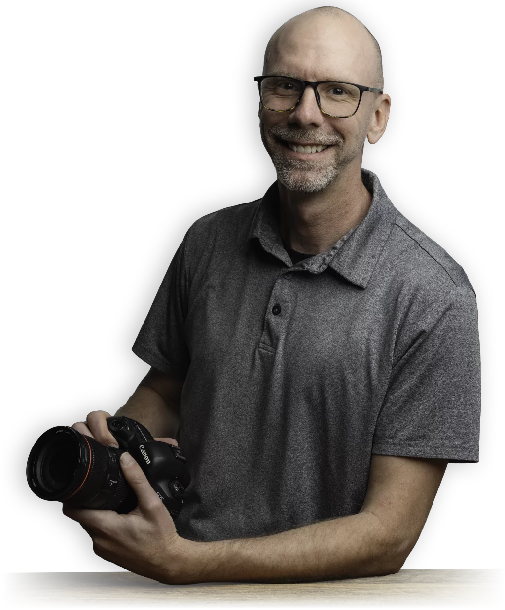 : Philip Meissner - Smiling bald man with glasses and a gray polo shirt holding a professional camera, sitting at a wooden table with a black background.