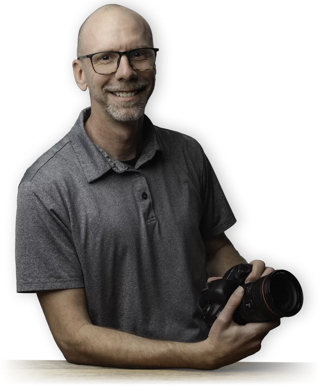 : Philip Meissner - Smiling bald man with glasses and a gray polo shirt holding a professional camera, sitting at a wooden table with a black background.