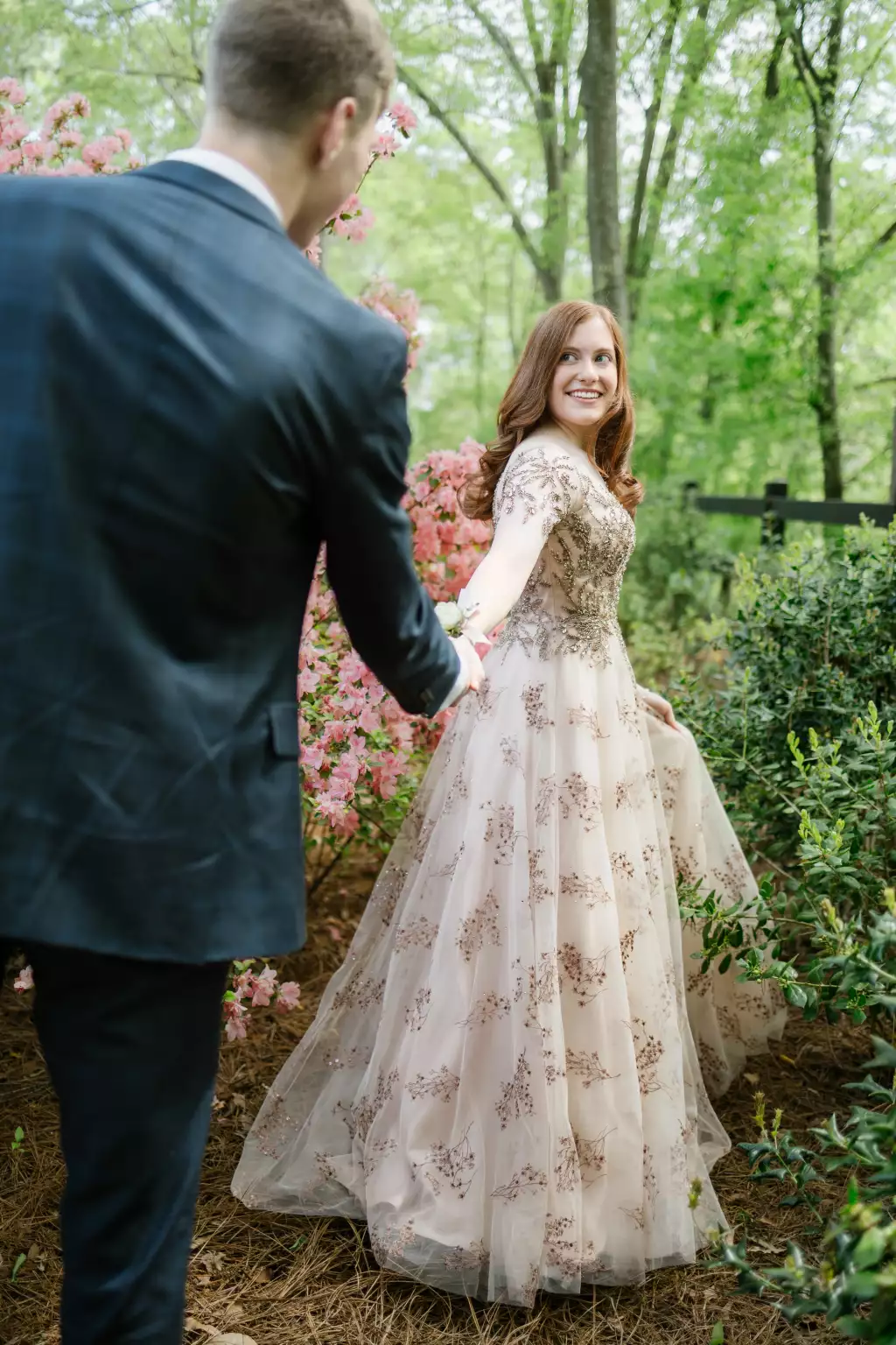 : A woman in an elegant, light-colored gown with embroidered floral details smiles while holding the hand of a man in a dark suit, leading him through a lush garden with pink flowers and green foliage.