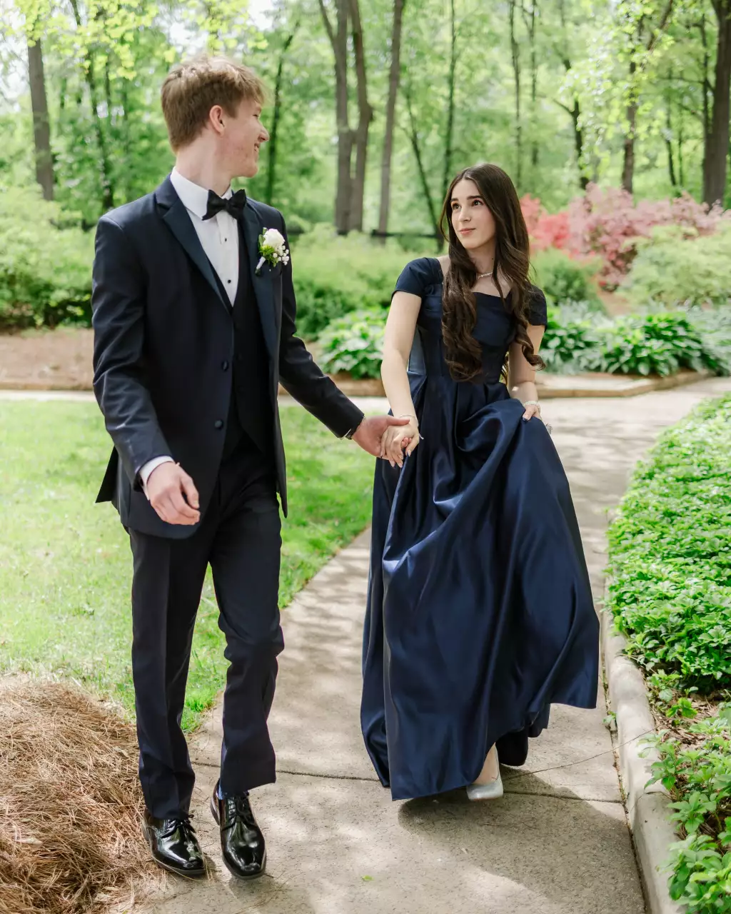 : Young couple dressed in formal attire walking hand in hand on a garden path surrounded by greenery, with the woman wearing a navy blue gown and the man in a dark suit with a bow tie and boutonniere.