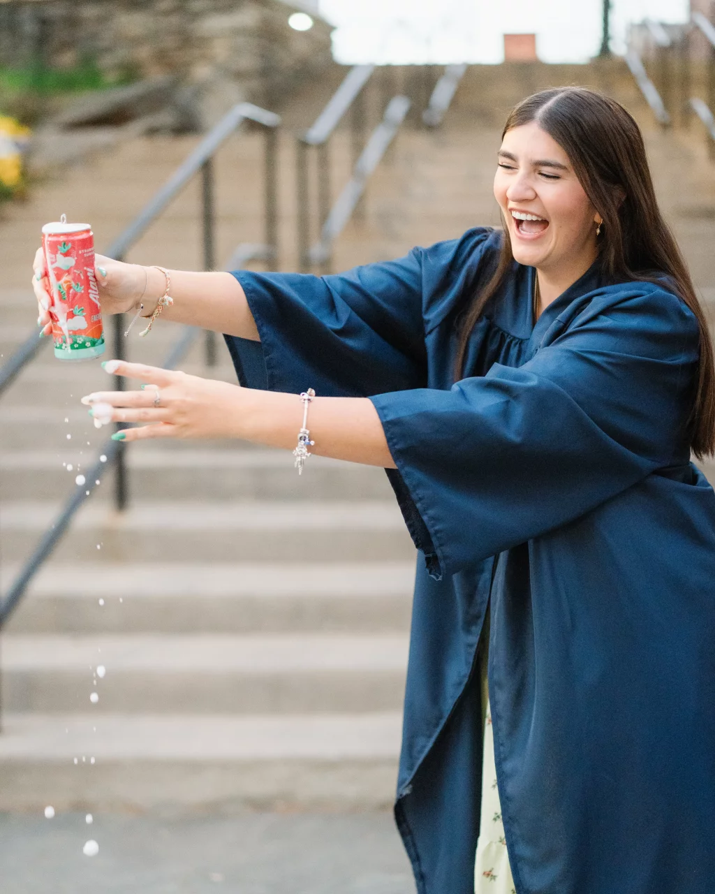 Alivia's senior photos taken Downtown, Greenville, SC: A young woman in a navy blue graduation gown laughs while pouring a drink from a red can, with liquid splashing out. She stands in front of outdoor steps.