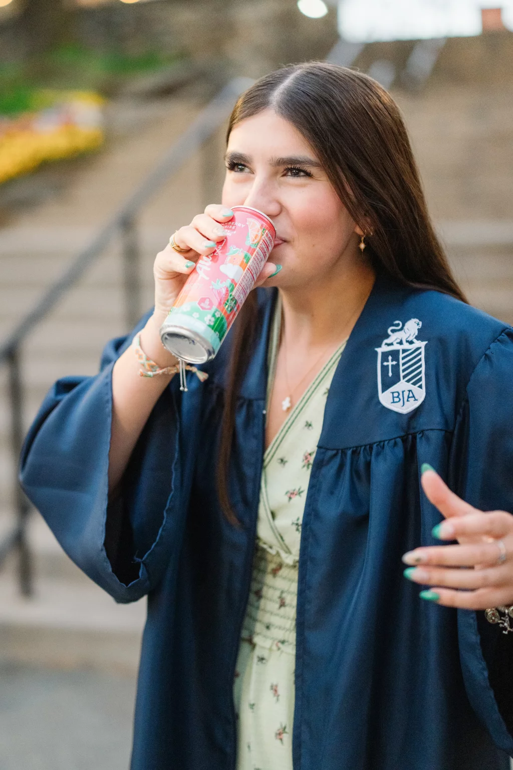 Alivia's senior photos taken Downtown, Greenville, SC: A young woman in a navy blue graduation gown drinks from a colorful can, standing outdoors in front of stone steps.