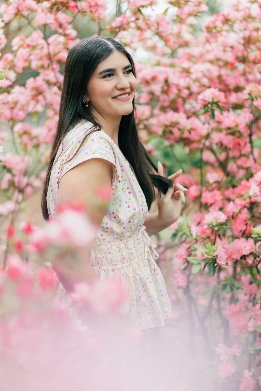 Alivia's senior photos taken Downtown, Greenville, SC: A young woman with long dark hair wearing a light floral dress stands smiling among blooming pink flowers.