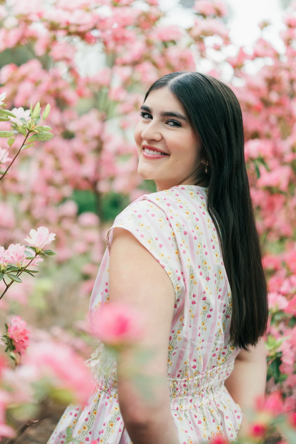Alivia's senior photos taken Downtown, Greenville, SC: A woman with long dark hair and a floral dress smiles over her shoulder, standing among blooming pink flowers.