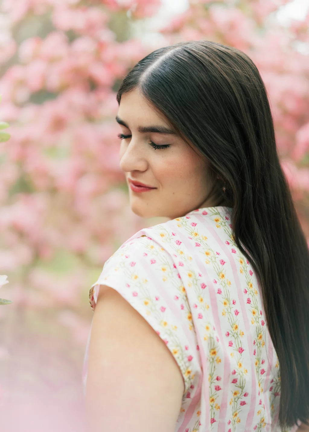Alivia's senior photos taken Downtown, Greenville, SC: A young woman with long brown hair stands outdoors with her eyes closed, wearing a light pink floral dress. The background is filled with soft-focus pink blossoms.