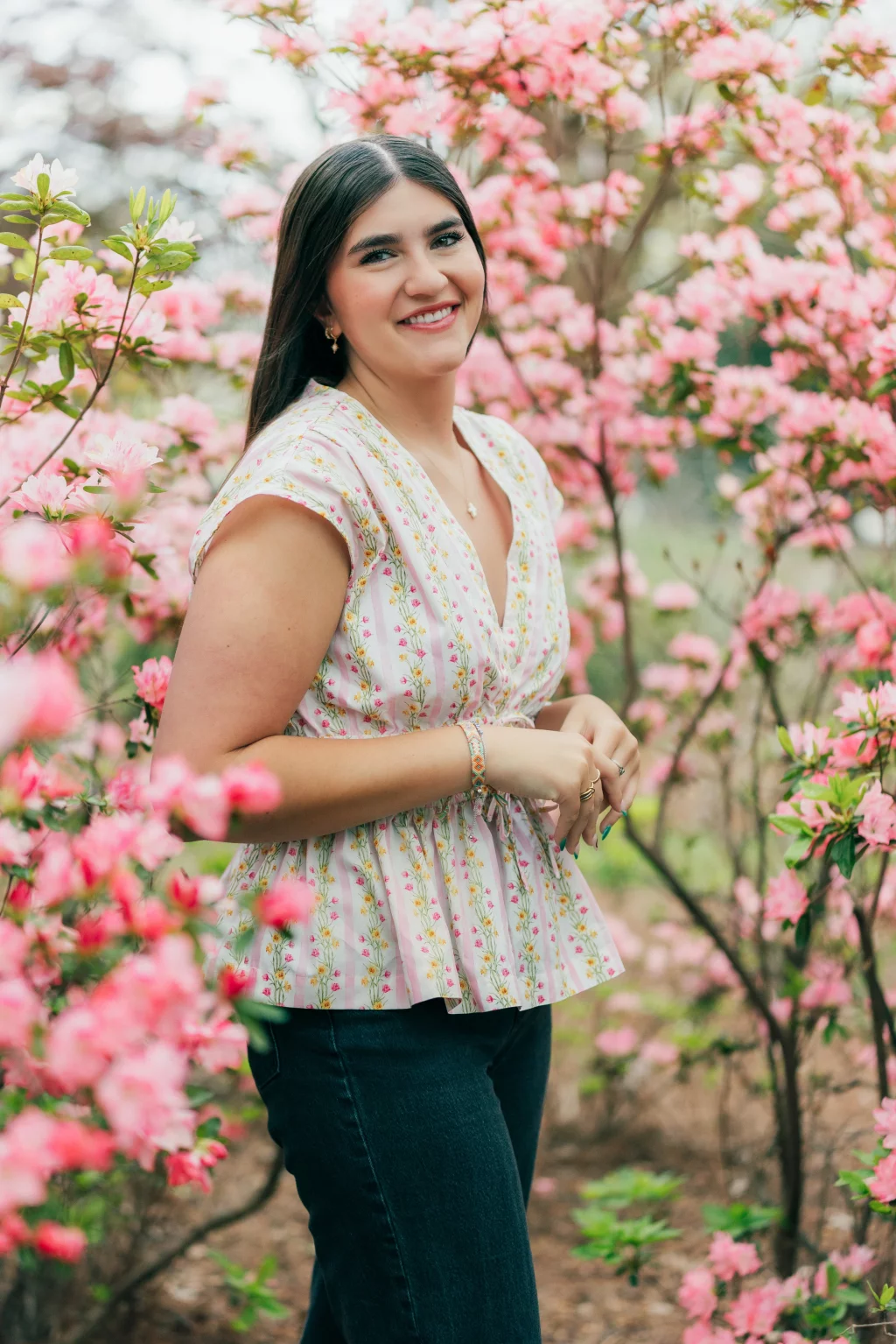 Alivia's senior photos taken Downtown, Greenville, SC: Woman with long dark hair wearing a floral blouse and dark jeans stands smiling among blooming pink flowers outdoors.