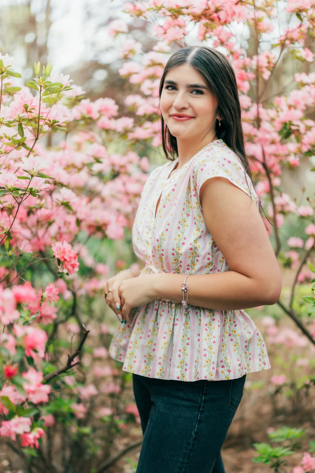 Alivia's senior photos taken Downtown, Greenville, SC: Woman with long brown hair stands among blooming pink flowers, wearing a white floral blouse and dark jeans, smiling at the camera.