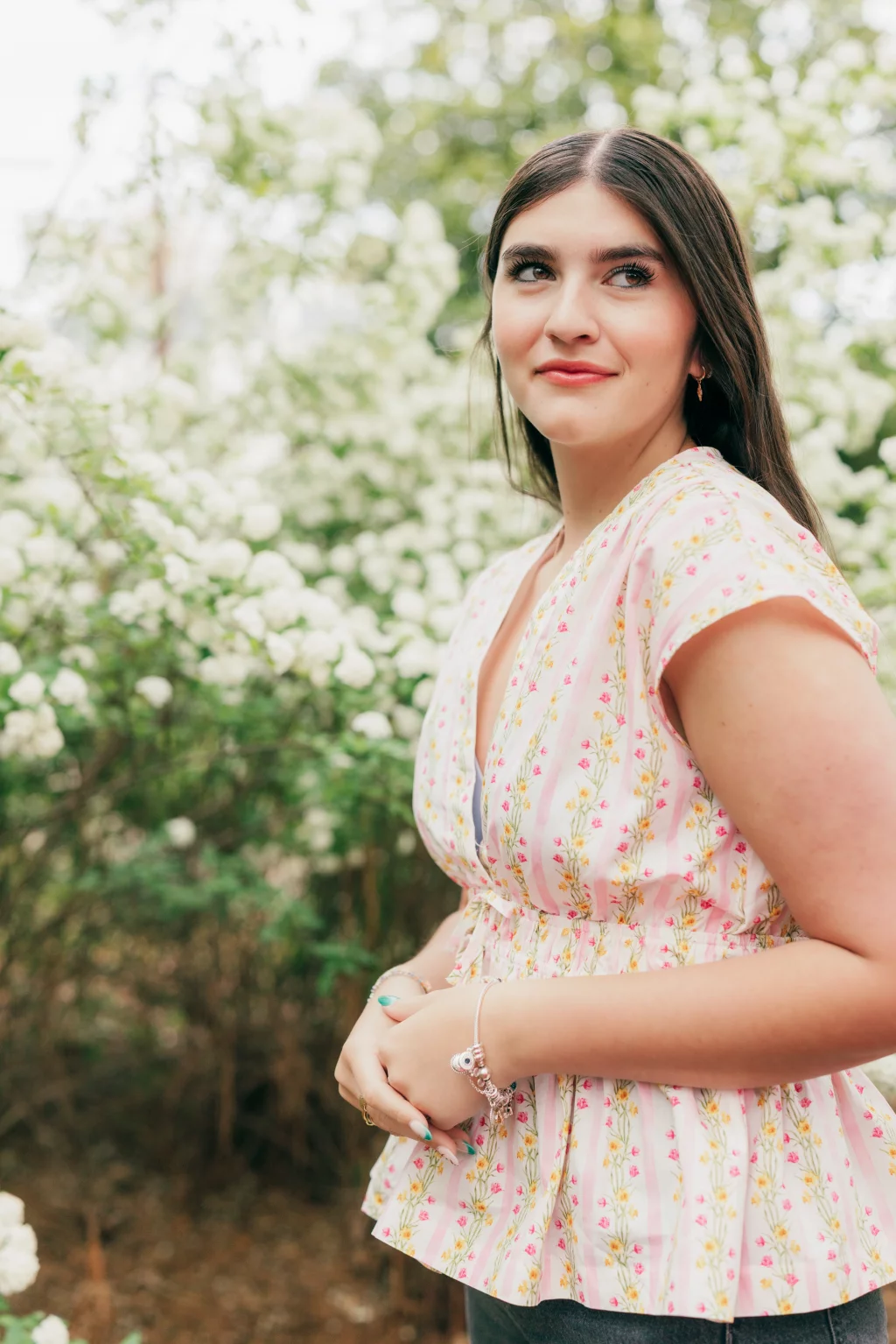 Alivia's senior photos taken Downtown, Greenville, SC: Young woman with long brown hair wearing a light pink floral blouse, standing outdoors in front of white flowering bushes, looking slightly to the side and smiling softly.