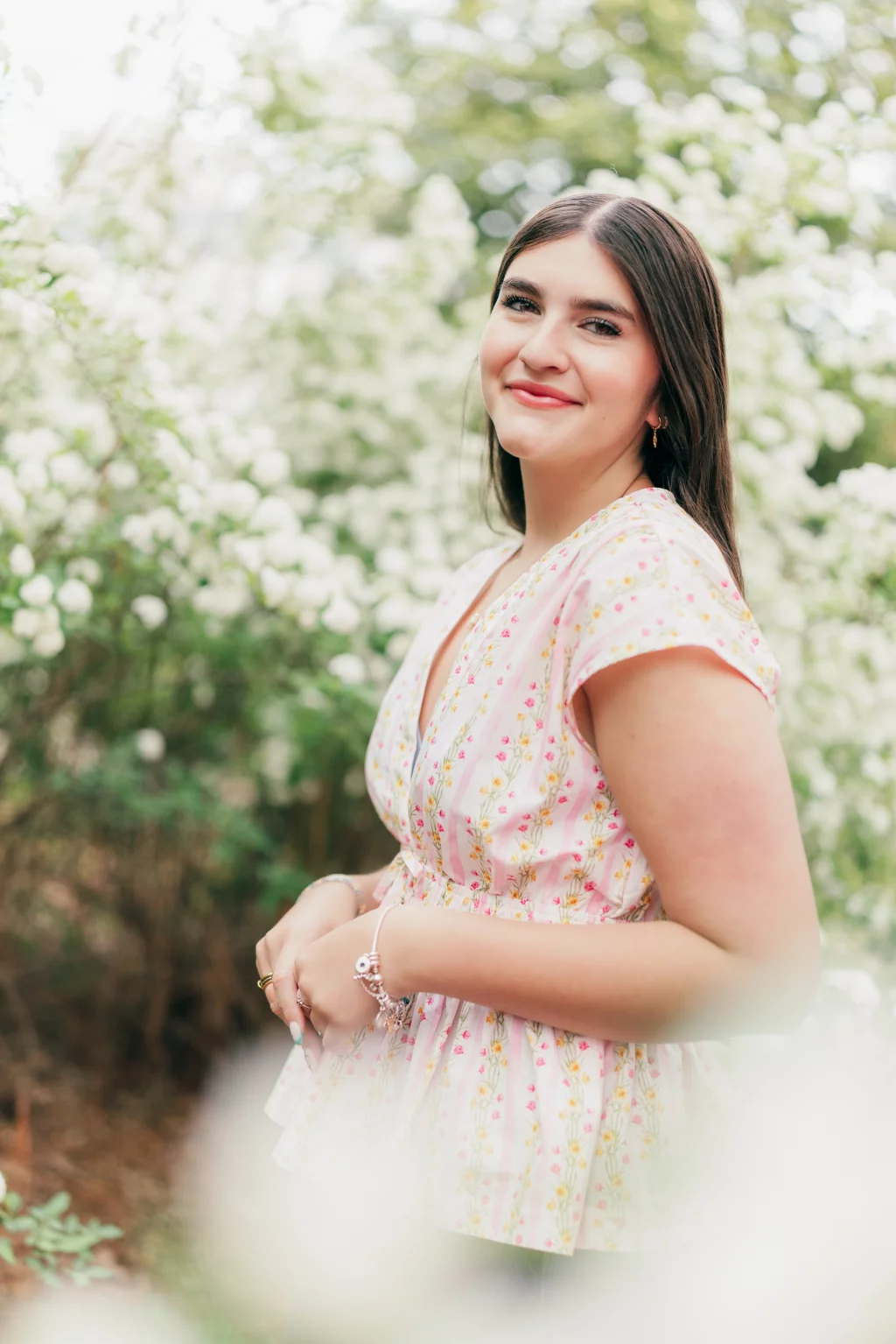 Alivia's senior photos taken Downtown, Greenville, SC: A young woman with long brown hair smiles while standing outdoors among blooming white flowers, wearing a light pink floral blouse.