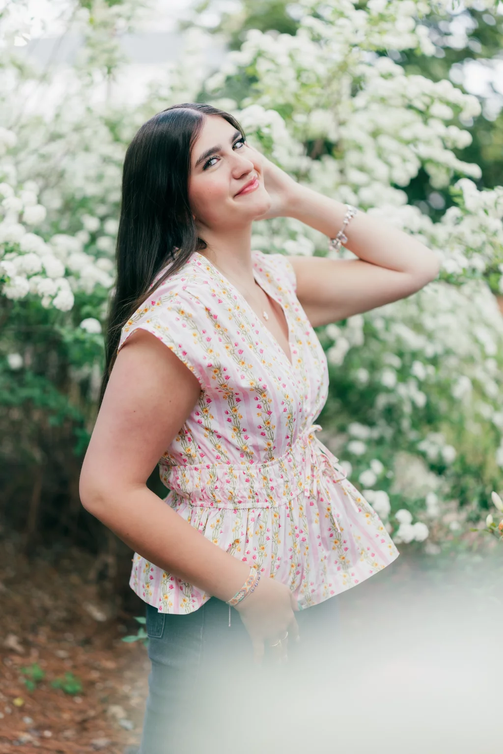 Alivia's senior photos taken Downtown, Greenville, SC: A young woman with long dark hair stands outdoors in front of blooming white flowers, wearing a light pink floral blouse and smiling with one hand touching her hair.
