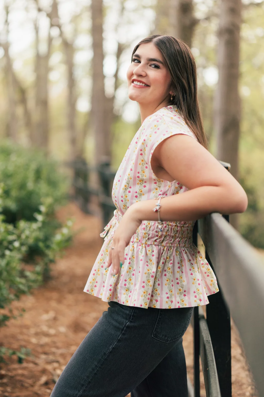 Alivia's senior photos taken Downtown, Greenville, SC: A young woman with long brown hair leans against a black fence outdoors, smiling at the camera. She is wearing a light pink floral blouse and dark jeans, with trees and greenery in the background.