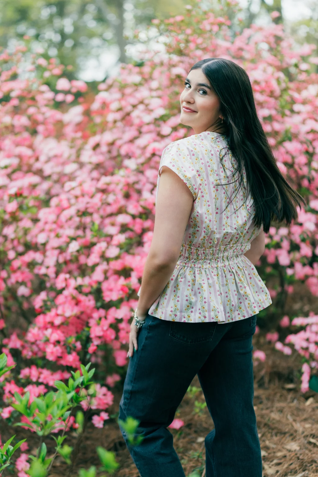 Alivia's senior photos taken Downtown, Greenville, SC: A woman with long dark hair stands outdoors in front of blooming pink flowers, wearing a light-colored, floral-patterned blouse and dark pants, looking over her shoulder and smiling gently at the camera.