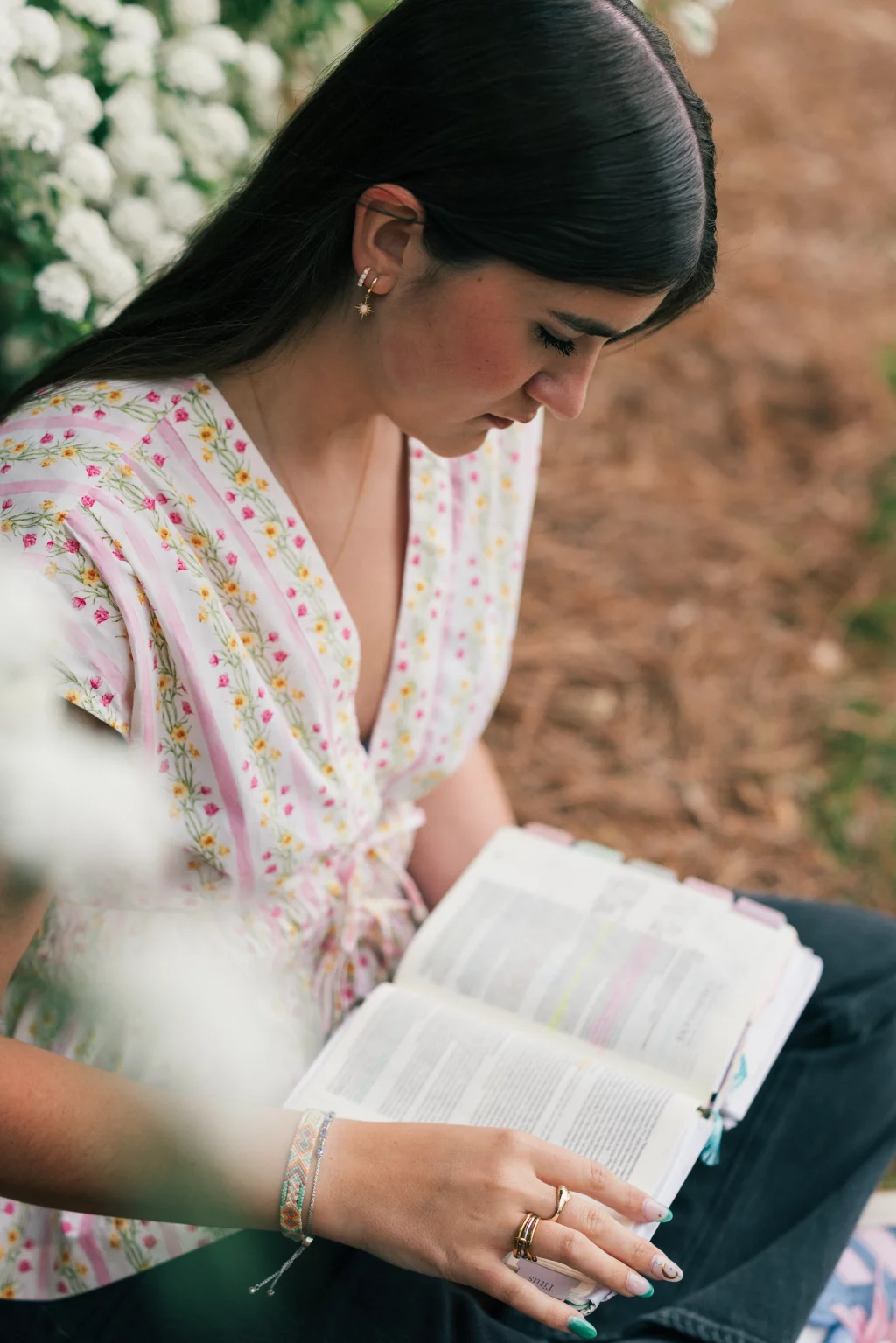 Alivia's senior photos taken Downtown, Greenville, SC: Young woman with long dark hair and floral blouse sitting outdoors, reading an open book with highlighted text, surrounded by white flowers and natural ground cover.