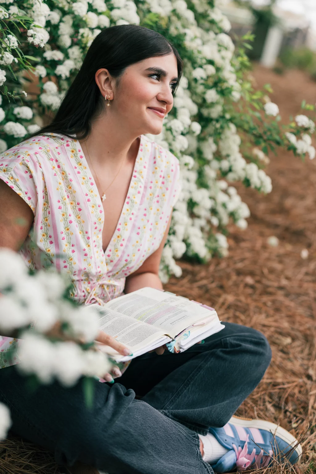 Alivia's senior photos taken Downtown, Greenville, SC: Young woman sitting outdoors beside white flowering bushes, smiling and holding an open book in her lap. She wears a floral blouse, dark jeans, and pastel sneakers.
