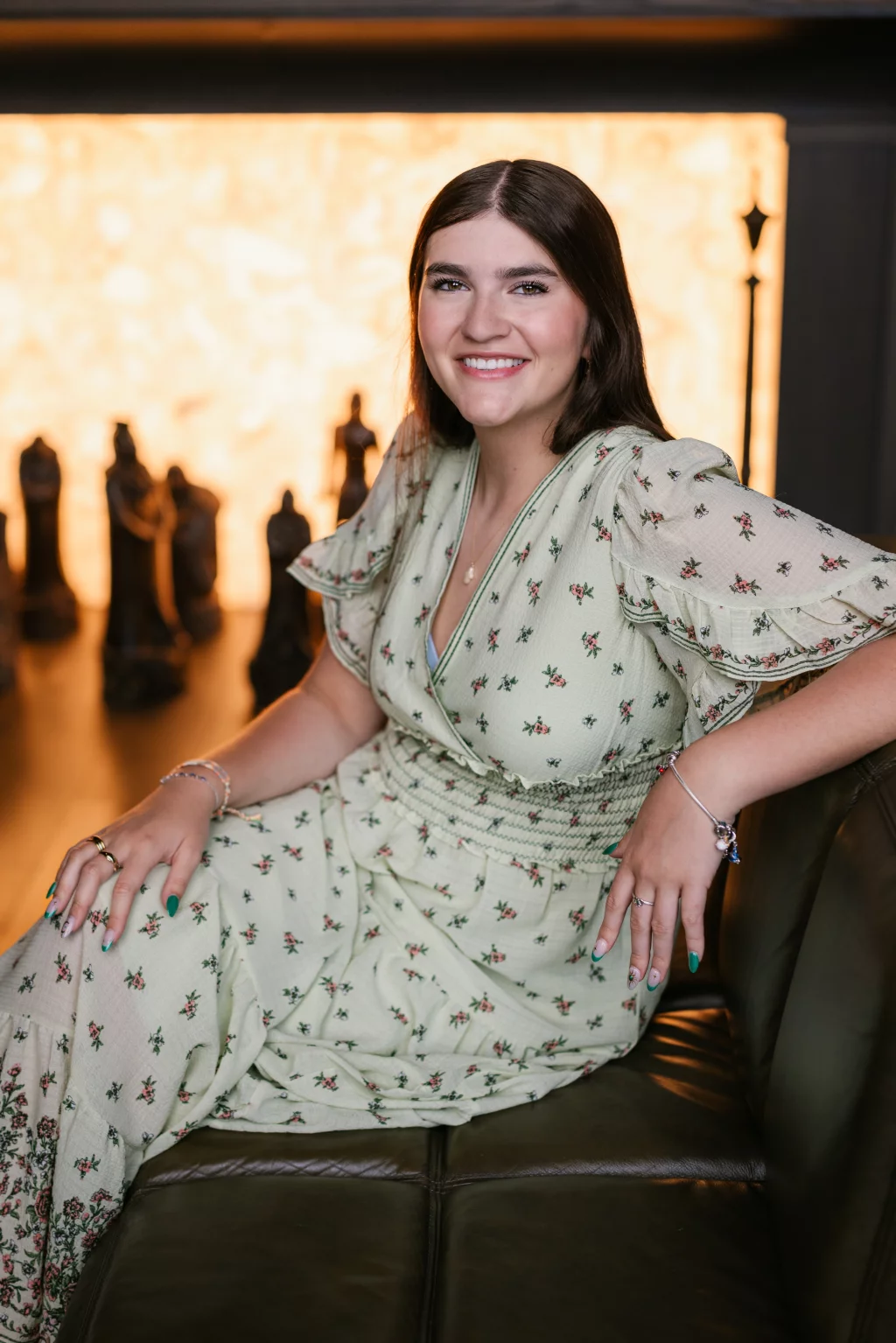 Alivia's senior photos taken Downtown, Greenville, SC: Woman with long brown hair wearing a light green floral dress, sitting on a couch and smiling at the camera, with blurred chess pieces and a warm, glowing background behind her.