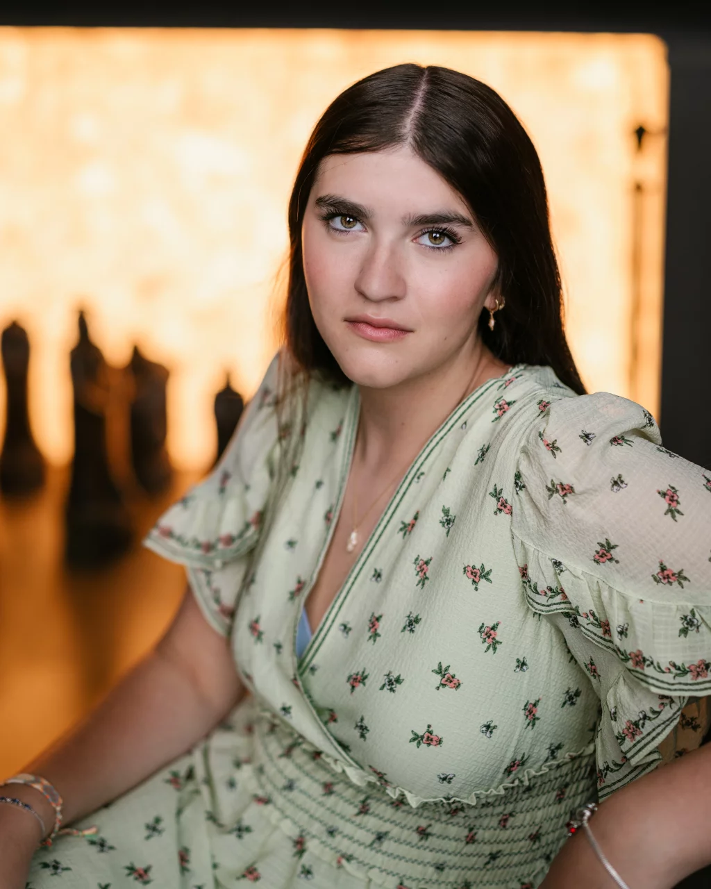 Alivia's senior photos taken Downtown, Greenville, SC: A young woman with long dark hair and light eyes sits in front of a softly lit background, wearing a light green floral dress. Large chess pieces are blurred in the background.