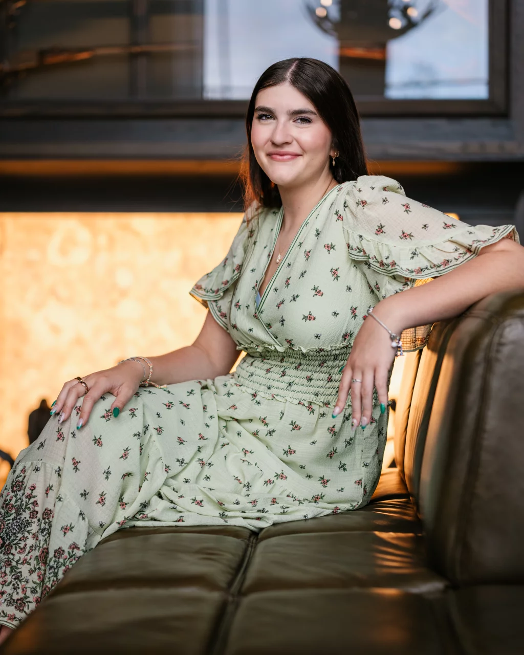Alivia's senior photos taken Downtown, Greenville, SC: A woman with long dark hair wearing a light green floral dress sits on a leather sofa, smiling at the camera. The background features a softly lit wall and a dark wooden shelf.