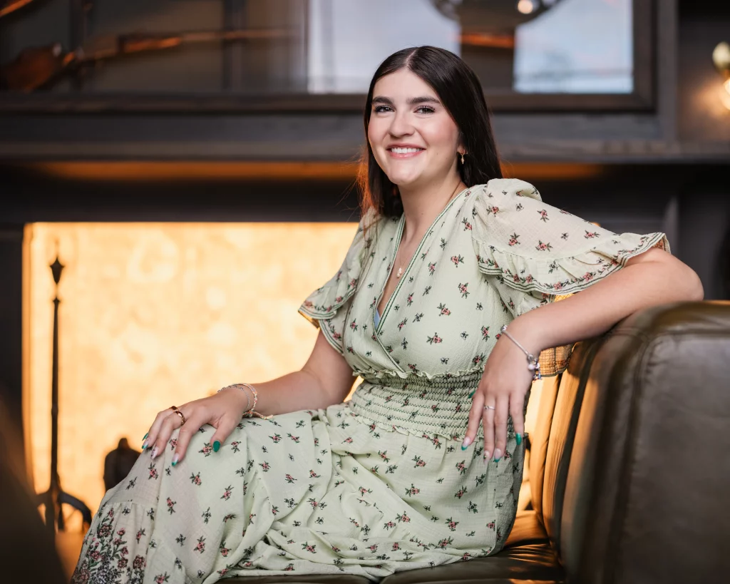 Alivia's senior photos taken Downtown, Greenville, SC: A woman with long dark hair, wearing a light green floral dress, sits on a leather couch and smiles at the camera in a warmly lit room.