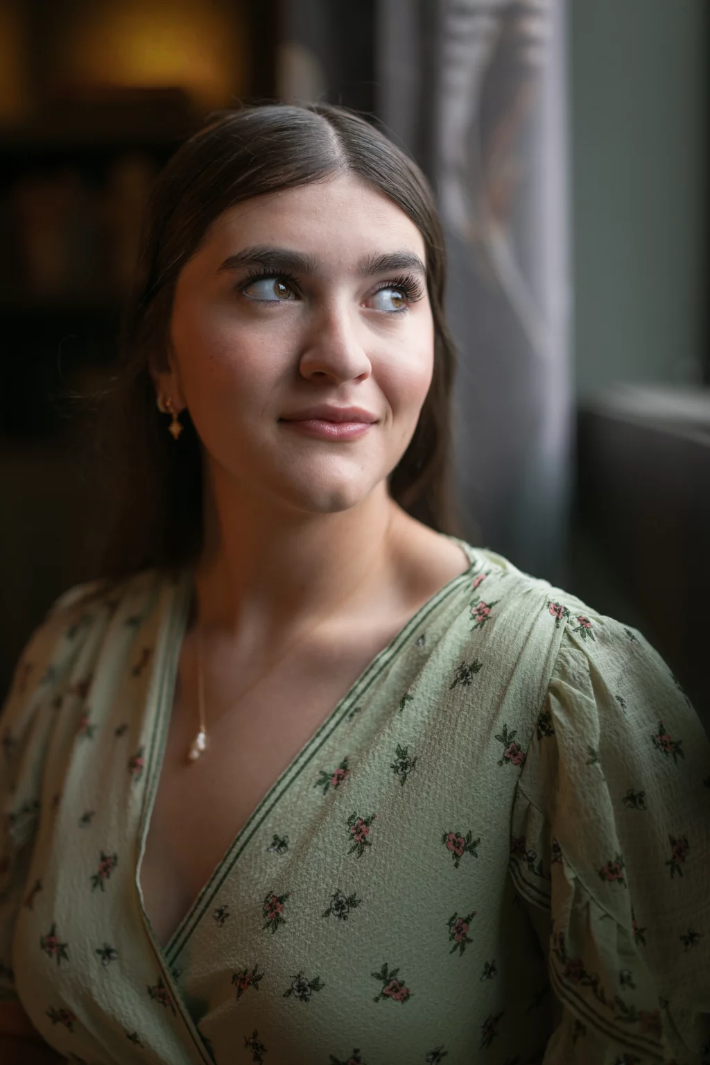 Alivia's senior photos taken Downtown, Greenville, SC: A young woman with long brown hair and light makeup looks out of a window, softly smiling. She is wearing a light green dress with a floral pattern and gold jewelry. The background is softly blurred.