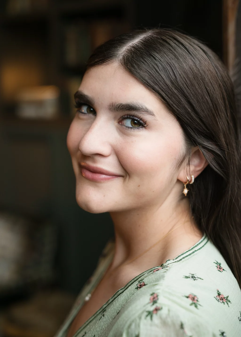 Alivia's senior photos taken Downtown, Greenville, SC: A young woman with long brown hair smiles softly at the camera. She is wearing a light green top with small floral patterns and gold hoop earrings with a star-shaped charm. The background is softly blurred.