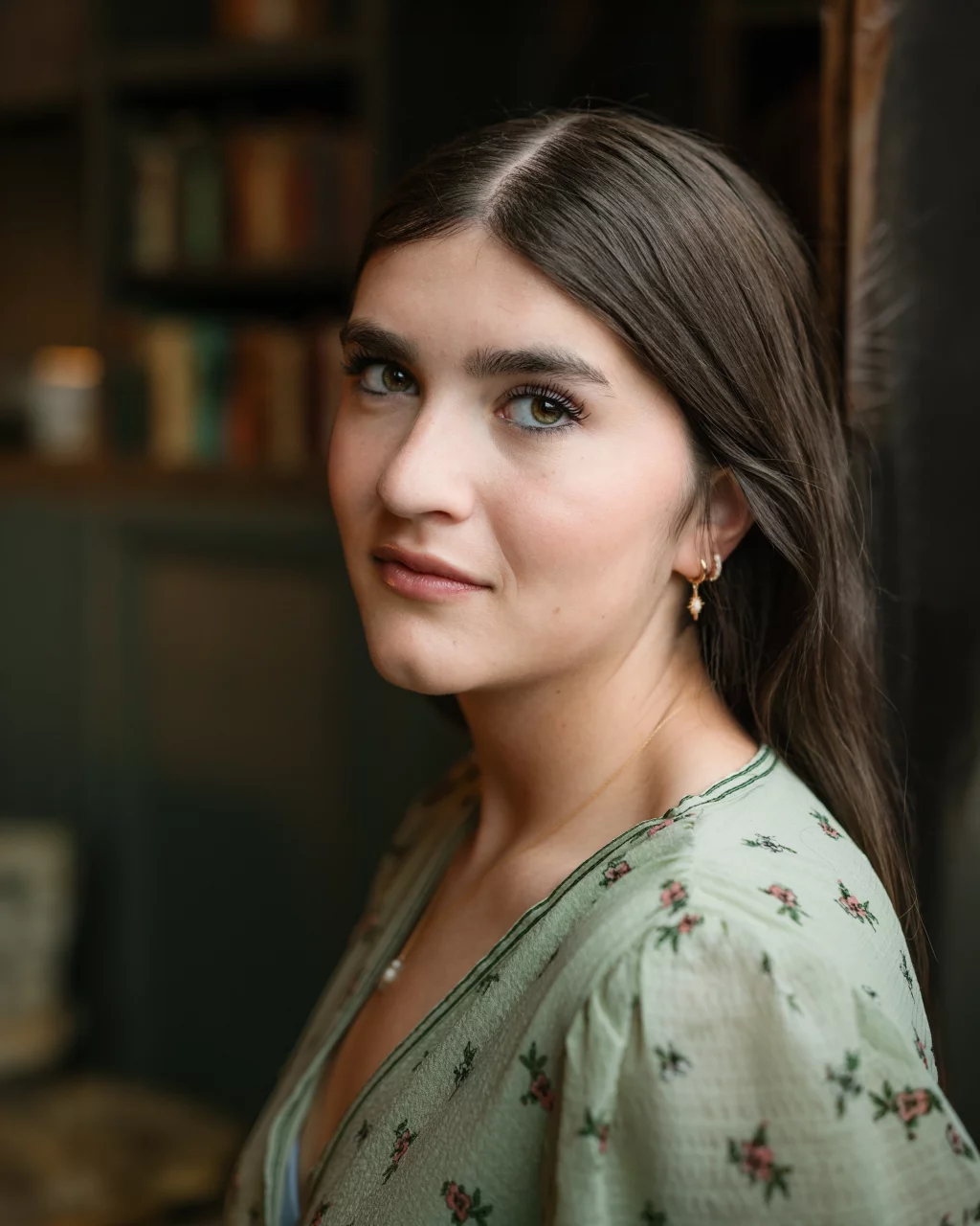 Alivia's senior photos taken Downtown, Greenville, SC: Young woman with long brown hair and light eyes, wearing a green floral blouse and gold earrings, looking at the camera with a soft expression in a softly lit indoor setting.