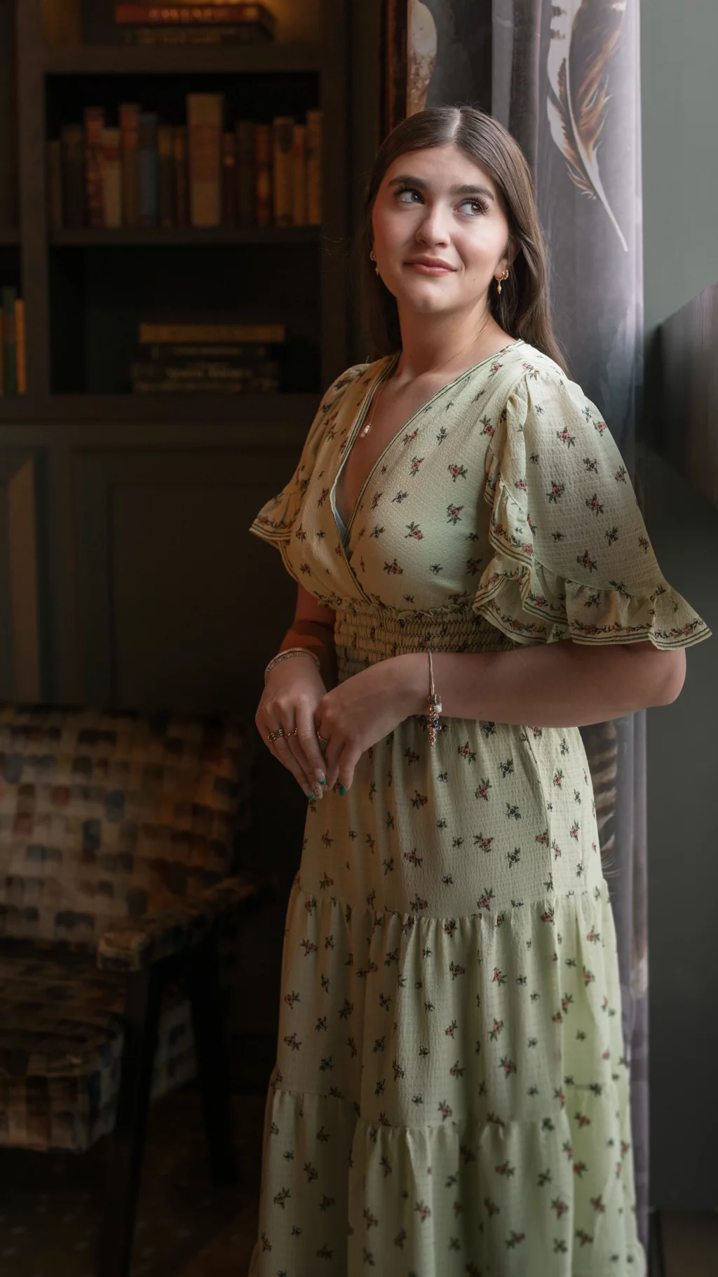 Alivia's senior photos taken Downtown, Greenville, SC: A woman in a light green, floral-patterned dress stands indoors near a window, looking slightly upward and smiling softly. There is a bookshelf and an armchair in the background.