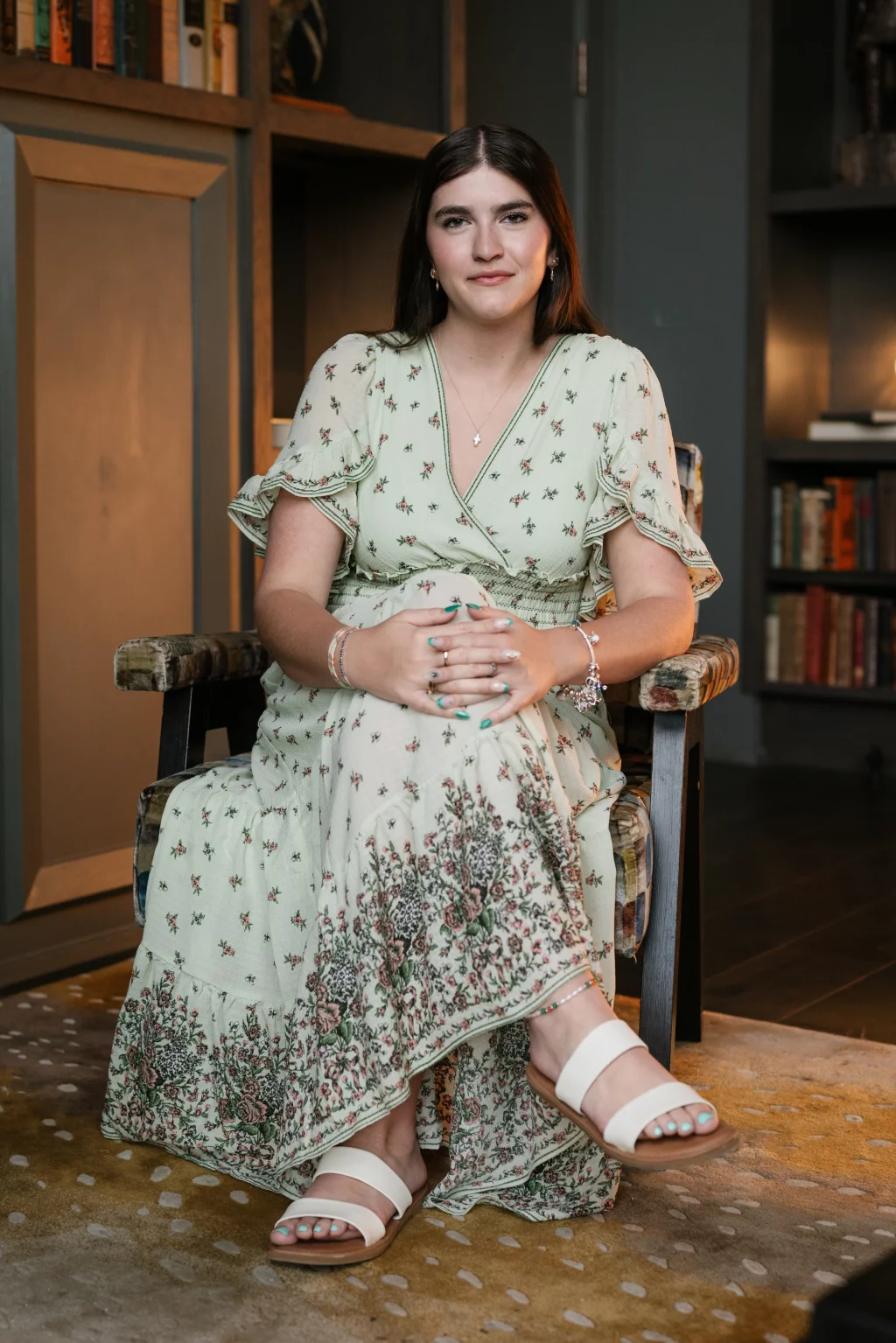 Alivia's senior photos taken Downtown, Greenville, SC: A woman with long brown hair sits in an armchair, wearing a light green floral dress and white sandals, with her hands folded in her lap. Bookshelves and a patterned rug are visible in the background.