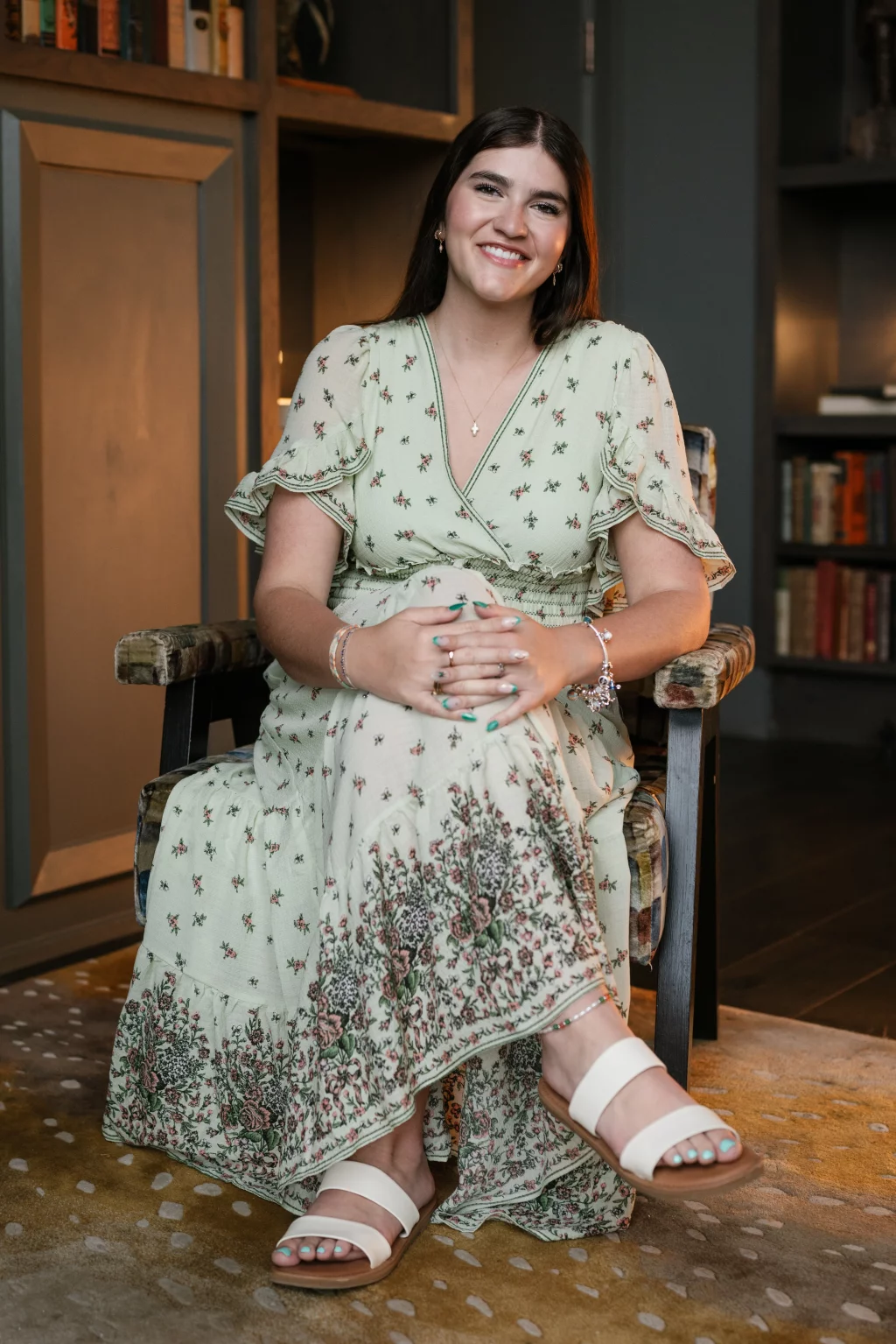 Alivia's senior photos taken Downtown, Greenville, SC: Young woman with long brown hair, wearing a light green floral dress and white sandals, sits smiling in an armchair with her hands folded in her lap; bookshelves are visible in the background.