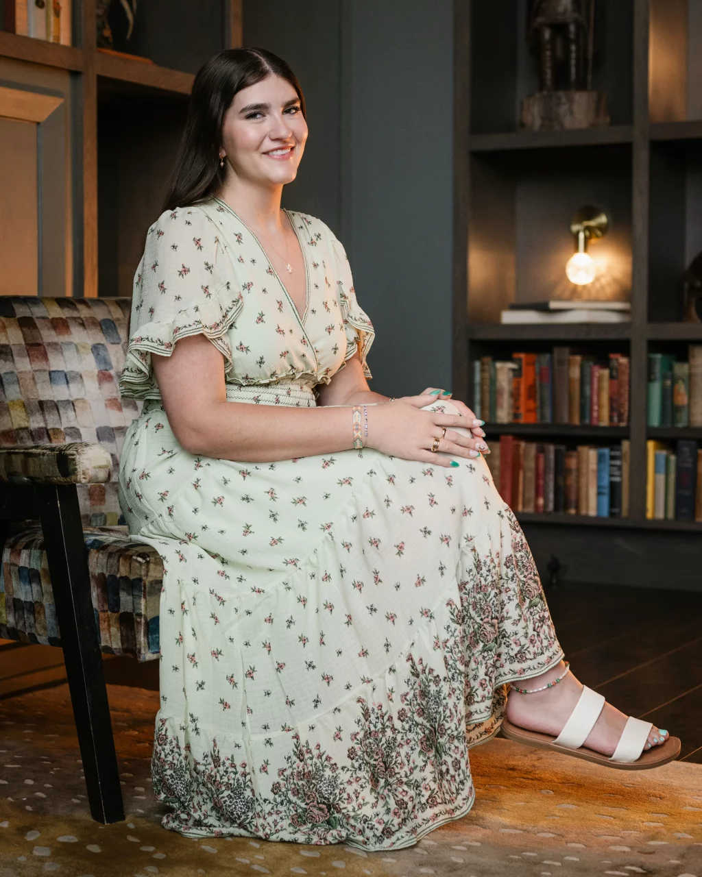 Alivia's senior photos taken Downtown, Greenville, SC: A woman in a light floral dress sits on a patterned chair, smiling, with her hands folded on her lap. Behind her is a bookshelf filled with books and a lit lamp.