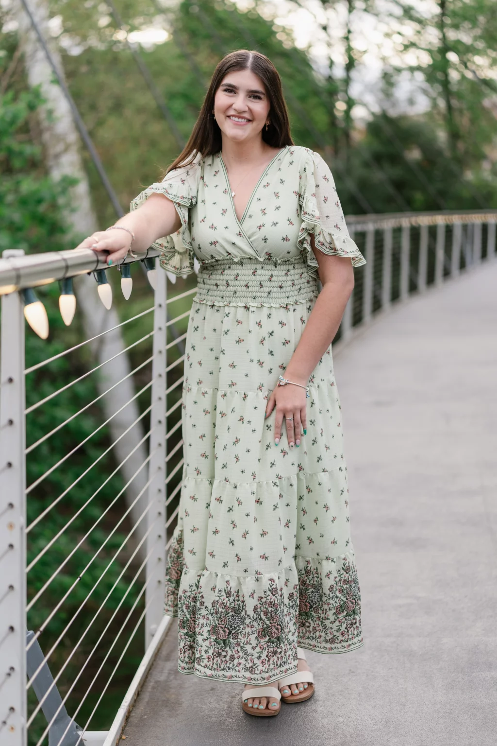 Alivia's senior photos taken Downtown, Greenville, SC: Young woman with long brown hair wearing a light green floral dress and sandals, smiling and standing on a pedestrian bridge, holding the railing decorated with string lights. Trees and greenery are visible in the background.