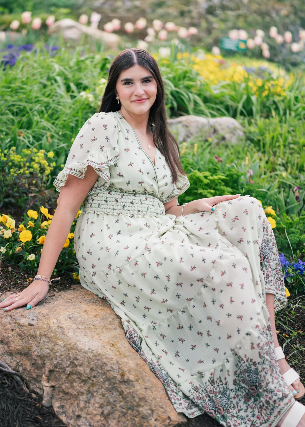 Alivia's senior photos taken Downtown, Greenville, SC: Young woman with long brown hair wearing a light green floral dress sits on a large rock in a garden with yellow and purple flowers and green foliage.