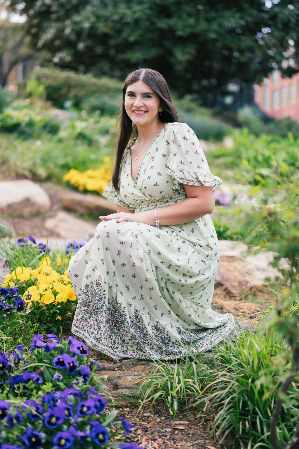 Alivia's senior photos taken Downtown, Greenville, SC: A woman with long brown hair in a light green floral dress kneels in a garden surrounded by purple and yellow flowers, smiling at the camera.