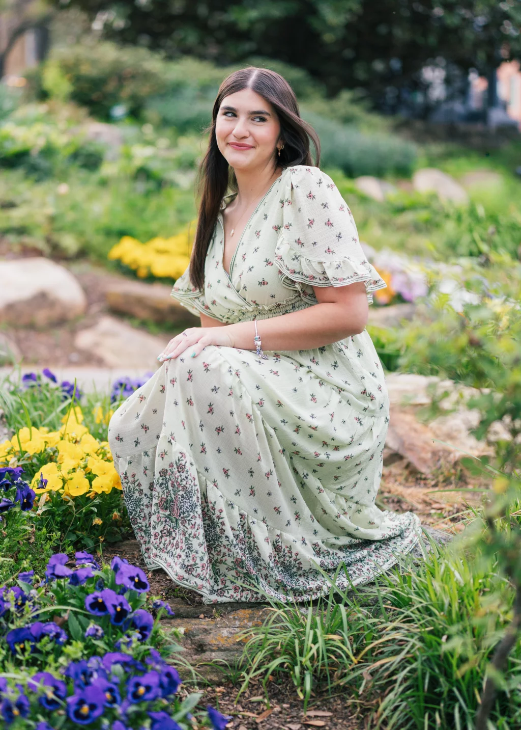 Alivia's senior photos taken Downtown, Greenville, SC: A woman in a light green floral dress kneels in a garden surrounded by yellow and purple flowers, smiling slightly and looking to the side.