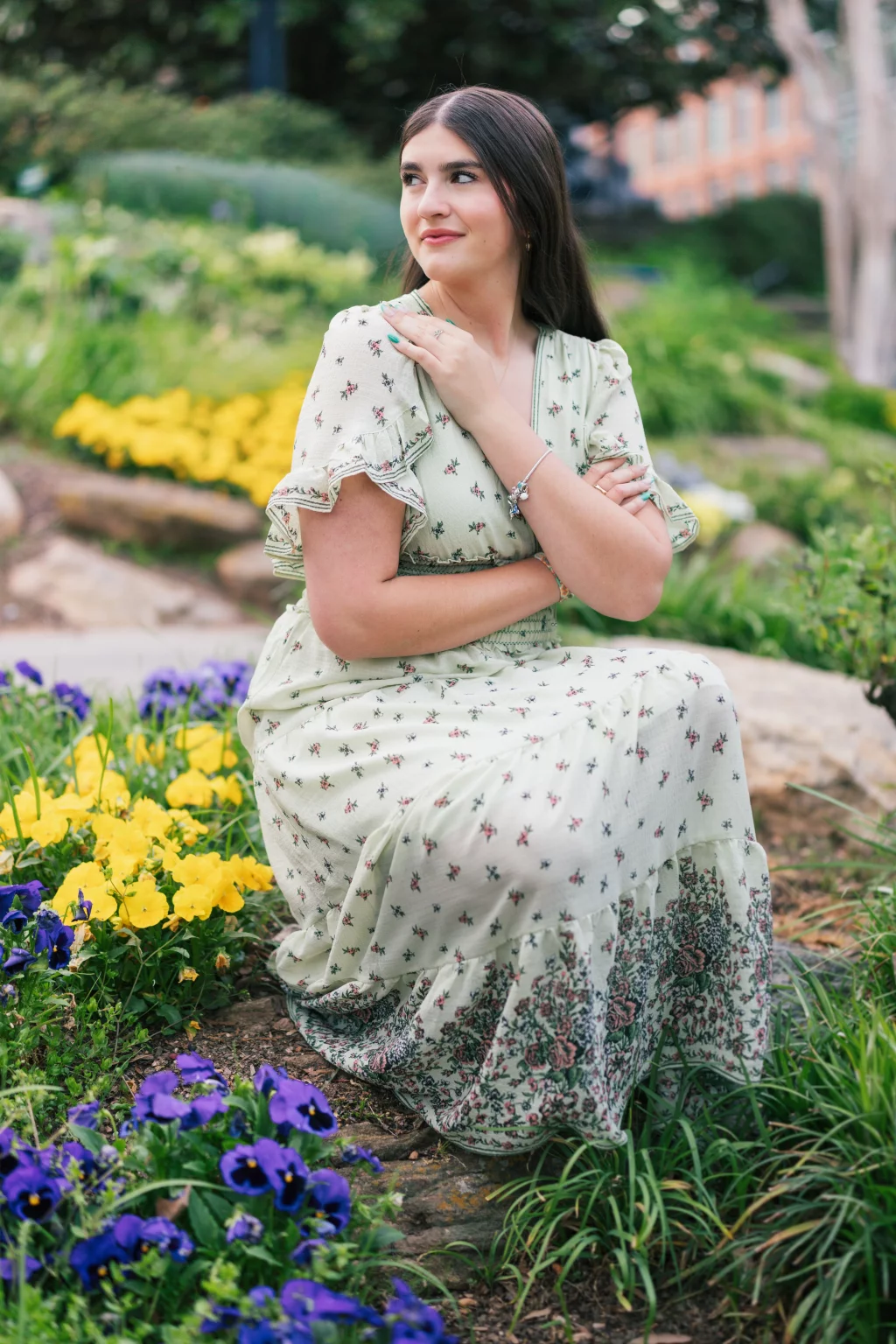 Alivia's senior photos taken Downtown, Greenville, SC: A young woman in a light floral dress kneels in a garden surrounded by purple and yellow flowers, looking to the side with a gentle smile.