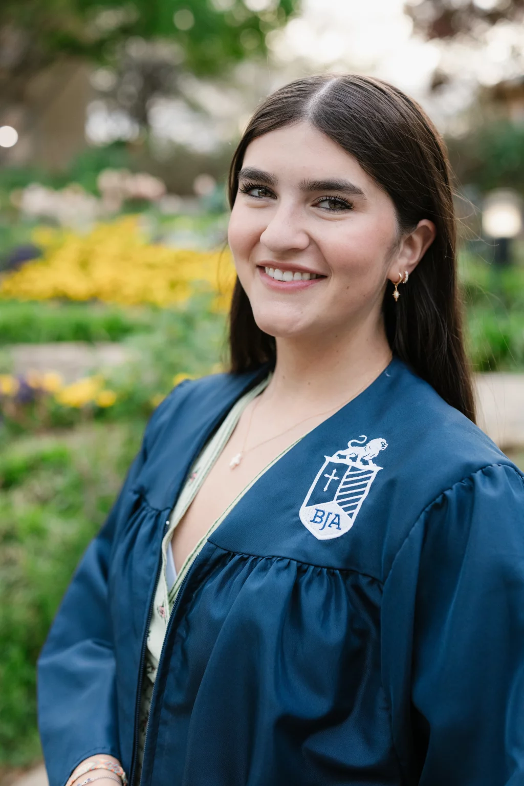 Alivia's senior photos taken Downtown, Greenville, SC: Young woman smiling outdoors, wearing a dark blue graduation gown with a white embroidered crest and the letters "BJA." Blurred background with greenery and yellow flowers.