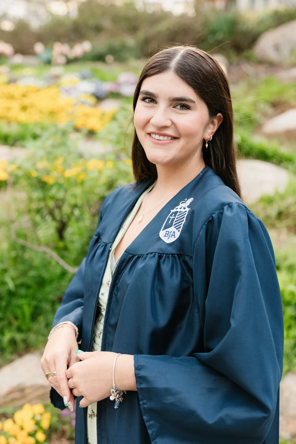Alivia's senior photos taken Downtown, Greenville, SC: Young woman smiling outdoors, wearing a dark blue graduation gown with a BJA crest, standing in front of yellow flowers and greenery.