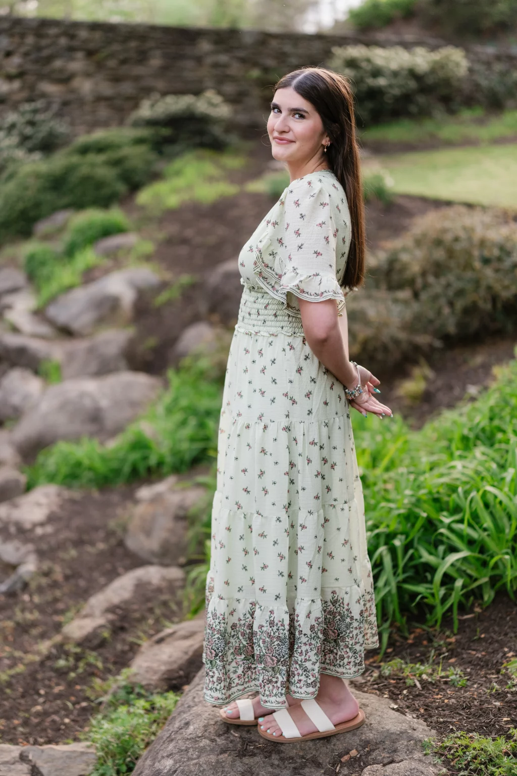 Alivia's senior photos taken Downtown, Greenville, SC: Woman with long brown hair in a light green floral dress stands outdoors on a rock, with greenery and a stone wall in the background. She is turned slightly to the side, hands clasped behind her back, and smiling at the camera.