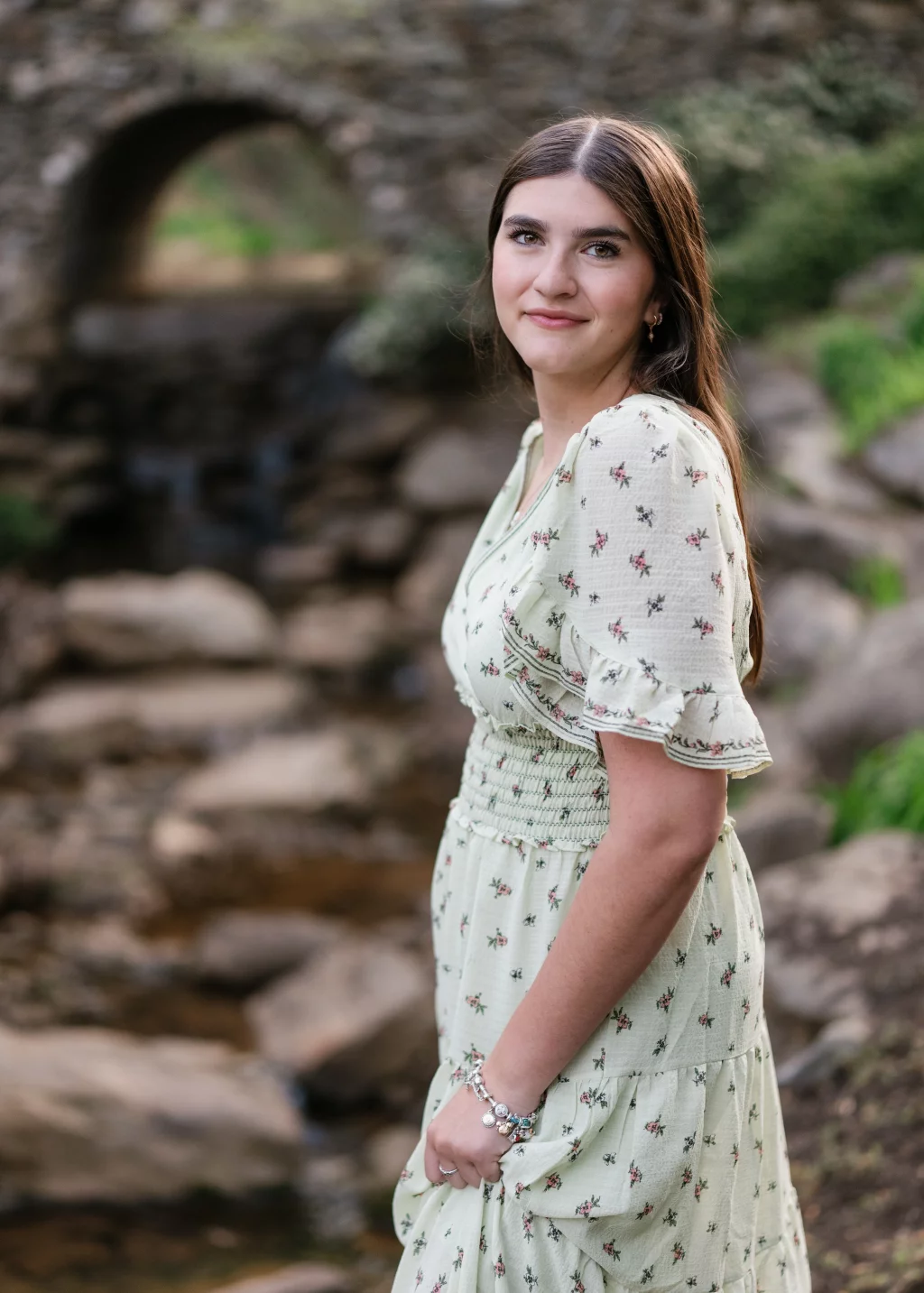Alivia's senior photos taken Downtown, Greenville, SC: Young woman with long brown hair wearing a light green floral dress, standing outdoors near a rocky stream with a stone arch bridge in the background.