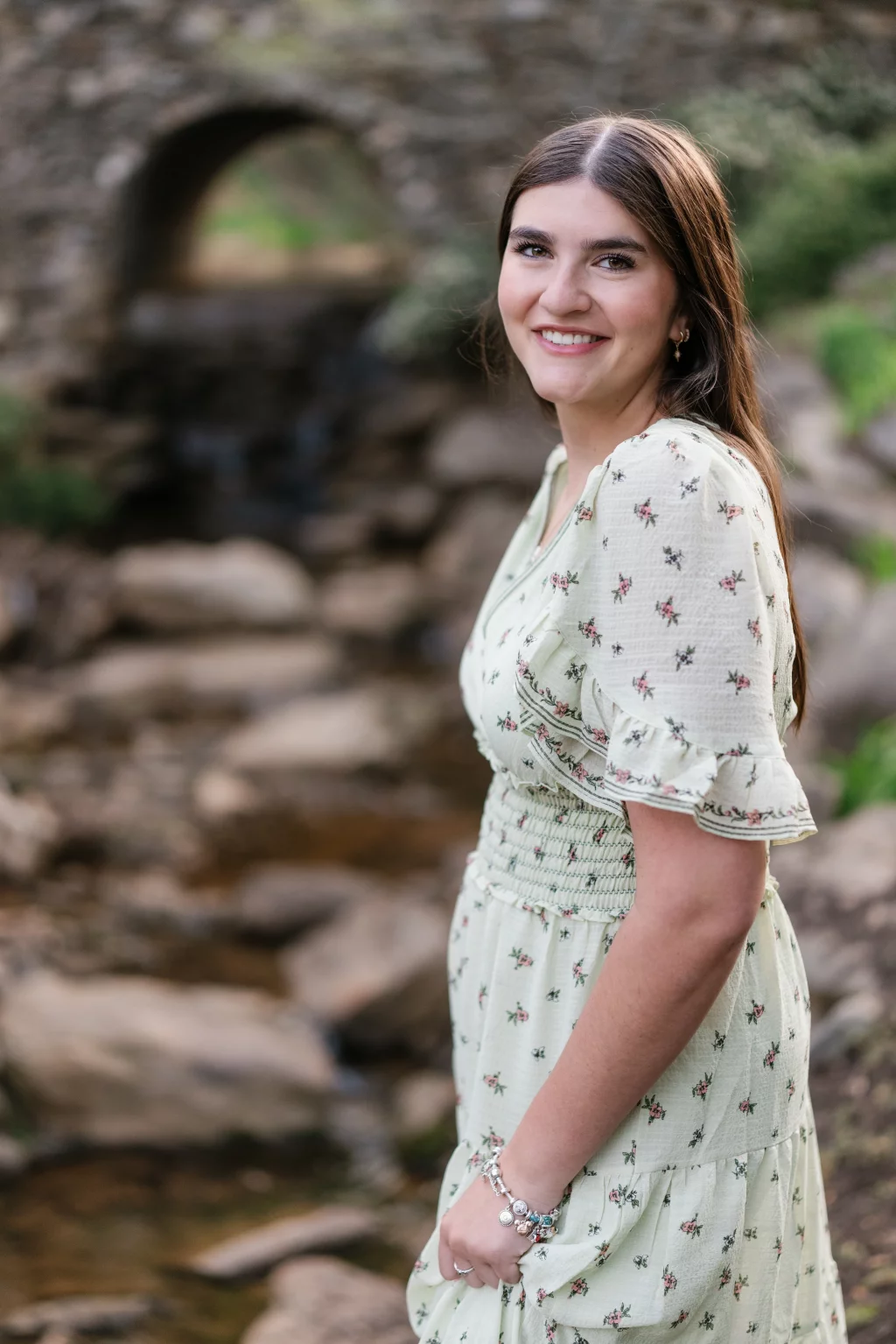 Alivia's senior photos taken Downtown, Greenville, SC: Young woman with long brown hair in a light green floral dress smiling outdoors, standing near a rocky creek with a stone bridge in the background.