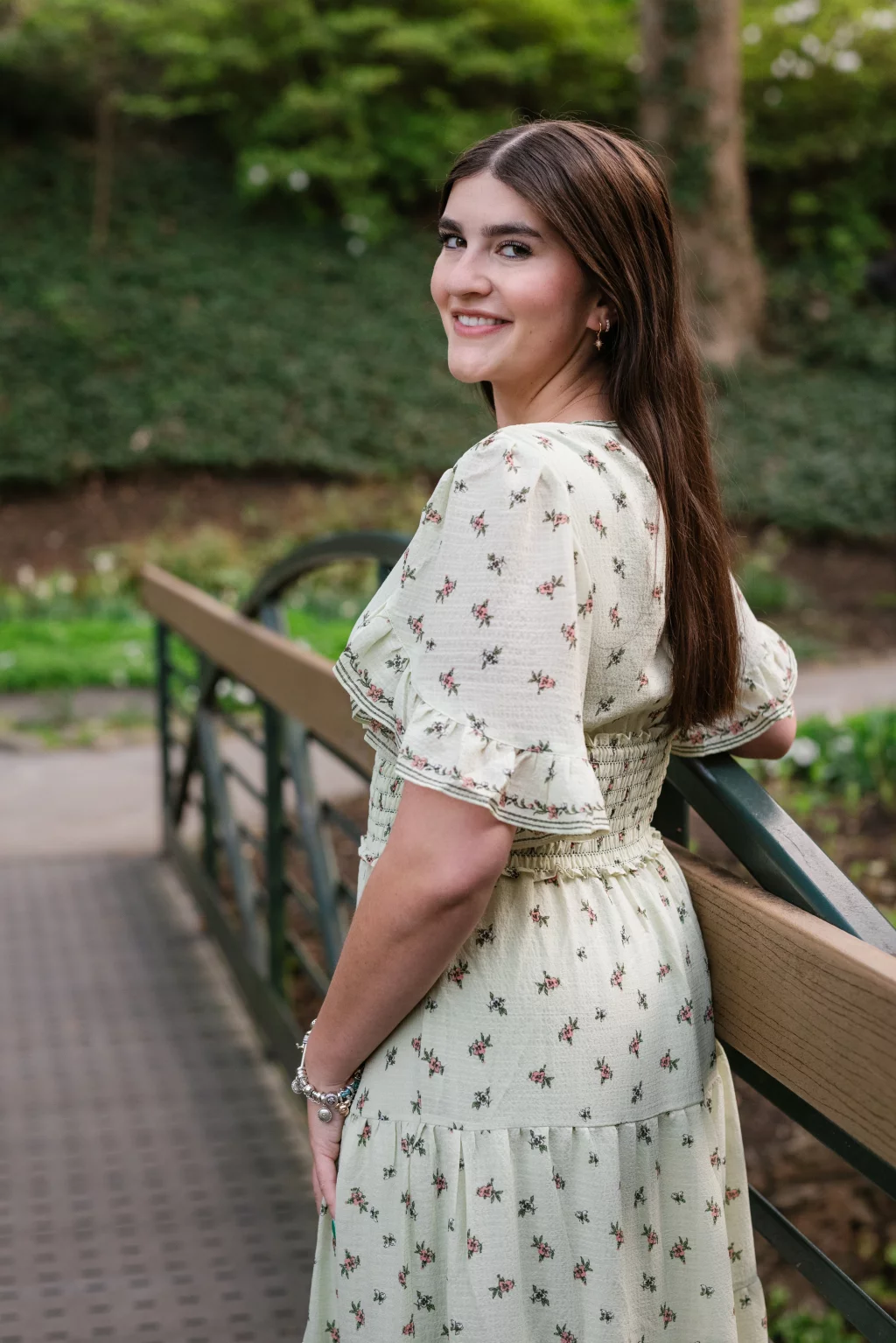 Alivia's senior photos taken Downtown, Greenville, SC: Woman with long brown hair in a light floral dress standing on a bridge, turned slightly and smiling at the camera, with greenery in the background.