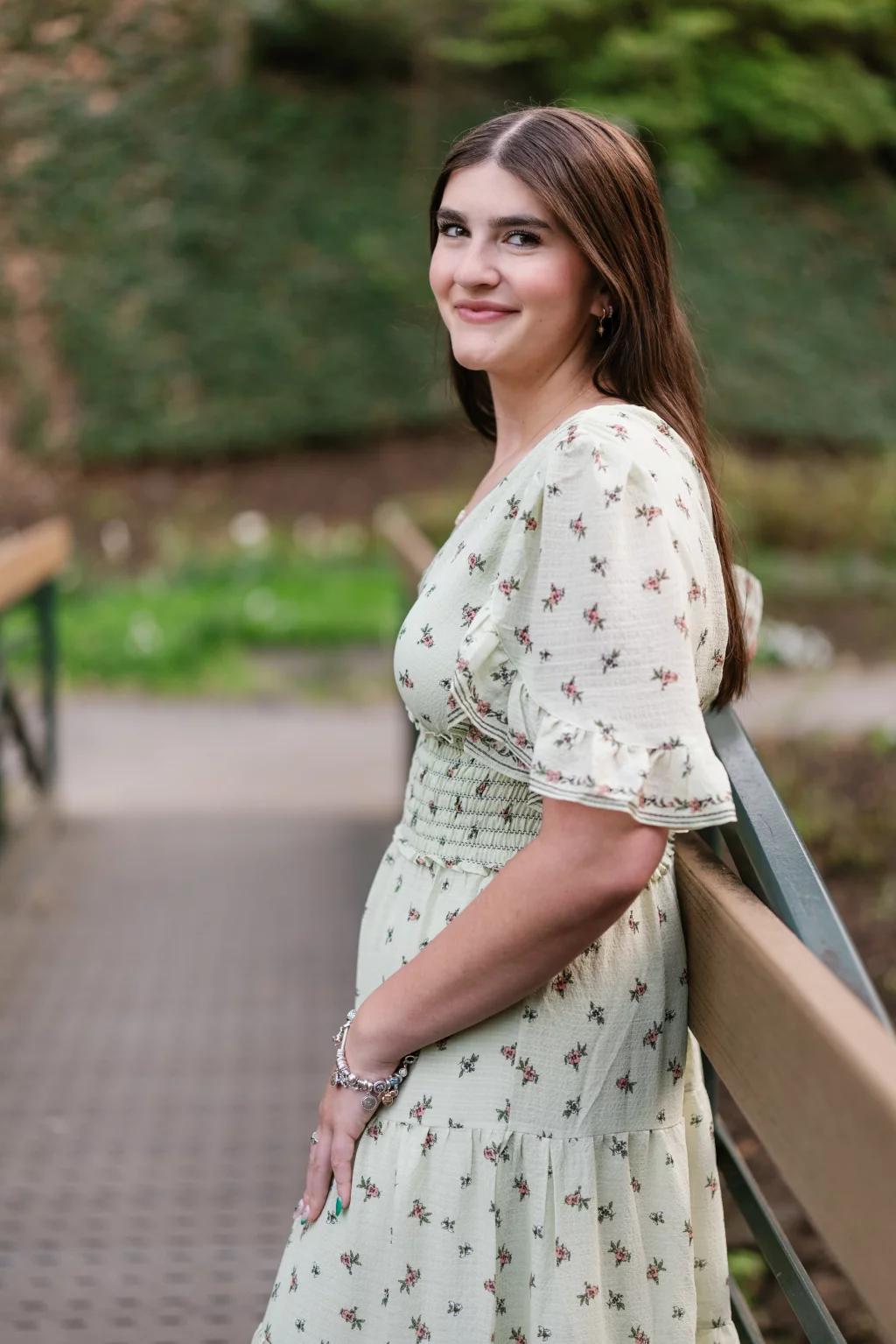 Alivia's senior photos taken Downtown, Greenville, SC: Young woman with long brown hair stands sideways on a bridge, wearing a light green floral dress, smiling at the camera with greenery in the background.