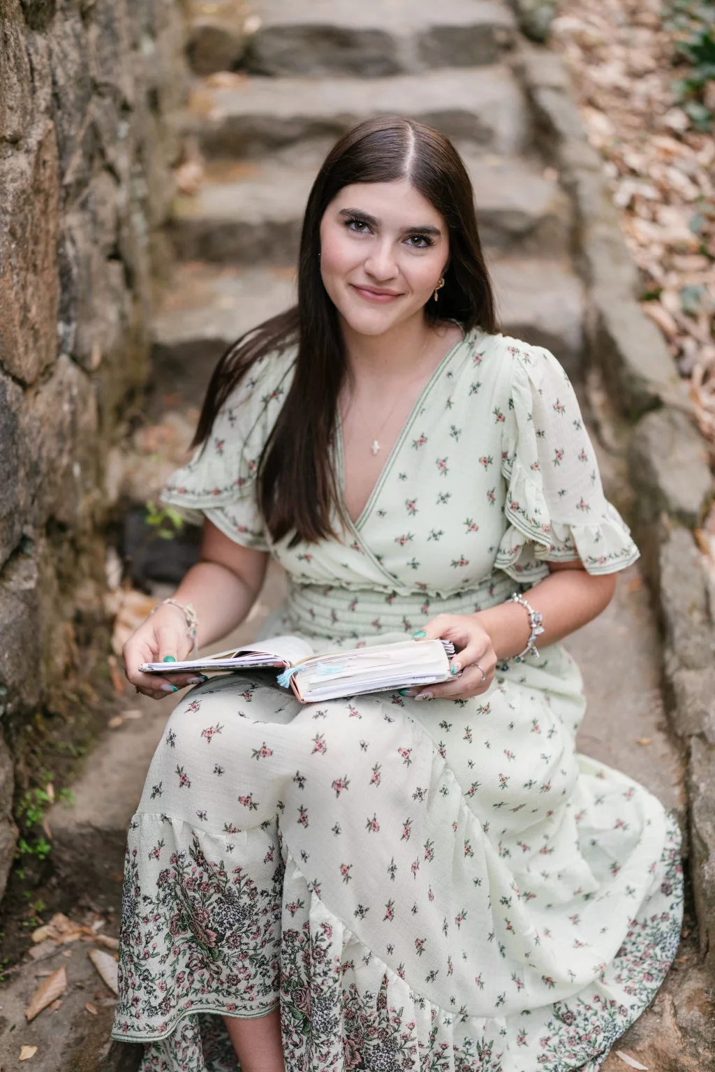 Alivia's senior photos taken Downtown, Greenville, SC: Young woman with long dark hair wearing a light green floral dress, sitting on stone steps outdoors, holding an open journal and smiling at the camera.