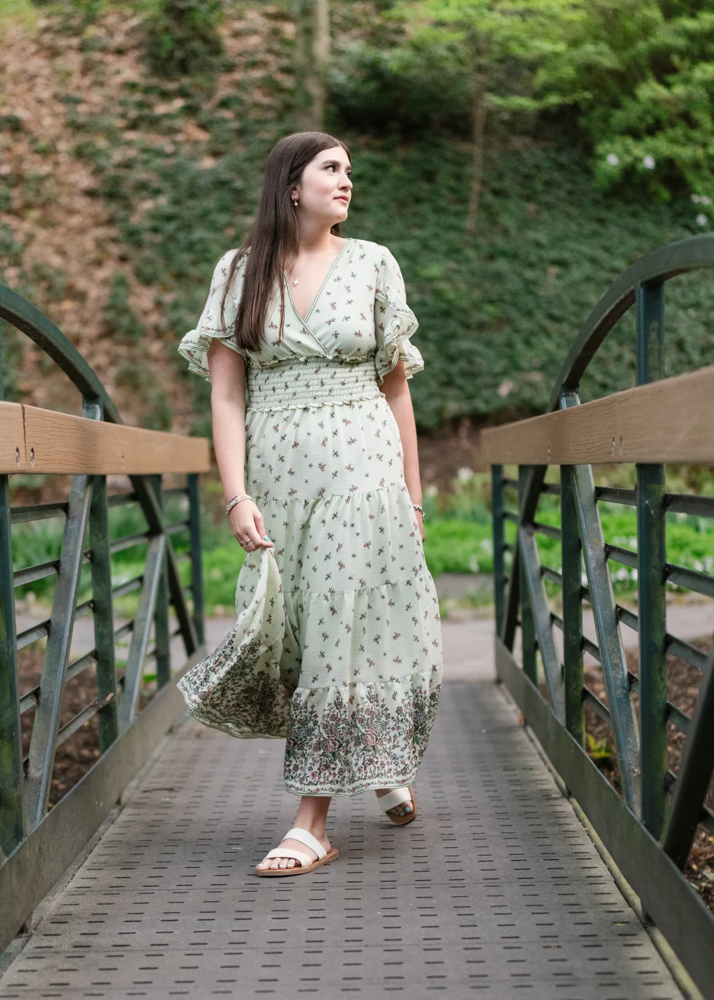 Alivia's senior photos taken Downtown, Greenville, SC: A woman in a light green floral dress walks across a metal footbridge, surrounded by greenery.
