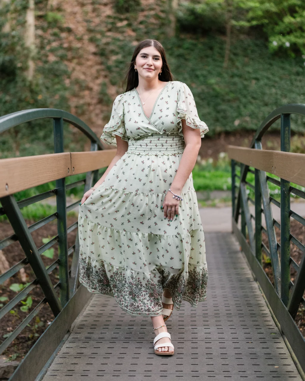 Alivia's senior photos taken Downtown, Greenville, SC: A woman in a light green floral dress stands on a metal footbridge outdoors, holding the skirt of her dress and smiling at the camera. Lush greenery and trees are visible in the background.