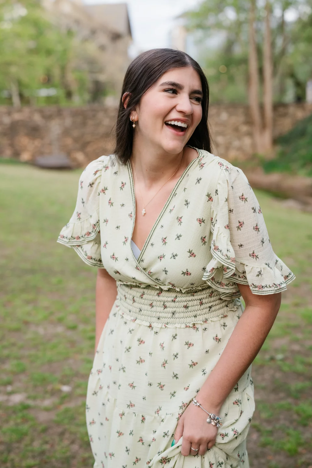 Alivia's senior photos taken Downtown, Greenville, SC: Young woman with long dark hair smiling outdoors, wearing a light floral dress with ruffled sleeves, standing on grass with trees and a stone wall in the background.