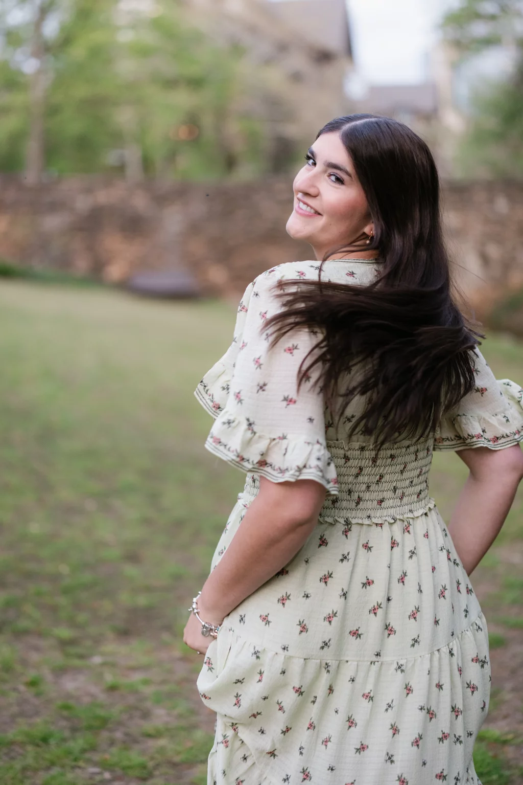 Alivia's senior photos taken Downtown, Greenville, SC: A young woman with long brown hair wearing a light-colored, floral-patterned dress stands outdoors, turning to smile at the camera. A stone wall and greenery are visible in the blurred background.