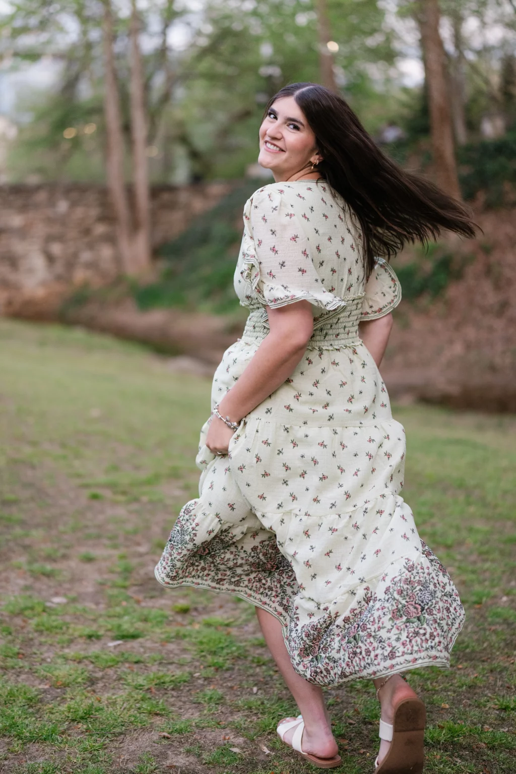 Alivia's senior photos taken Downtown, Greenville, SC: A young woman in a flowing, floral-print dress turns and smiles over her shoulder while walking outdoors on a grassy area, with trees and a stone wall in the background.