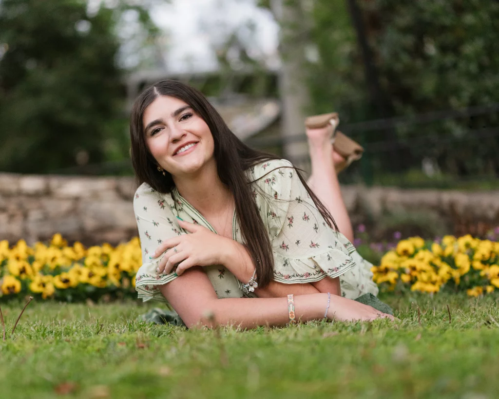 Alivia's senior photos taken Downtown, Greenville, SC: Young woman with long brown hair lying on grass, smiling, with yellow flowers and greenery in the background.