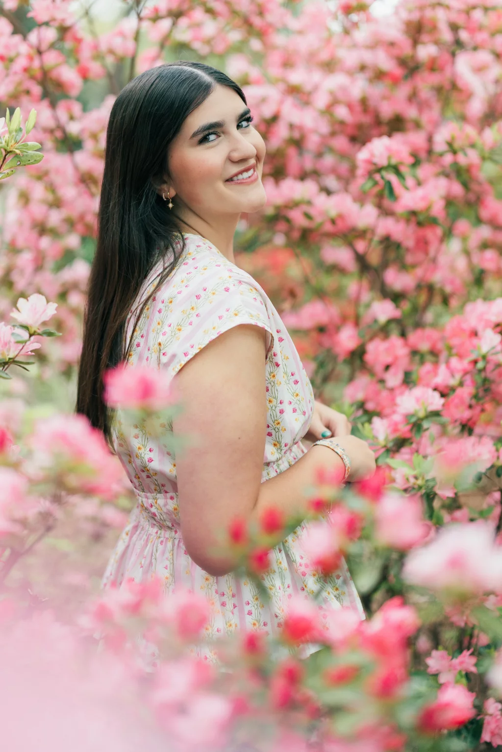Alivia's senior photos taken in downtown Greenville, SC: Woman with long dark hair in a light floral dress standing among bright pink blooming flowers, smiling over her shoulder at the camera.