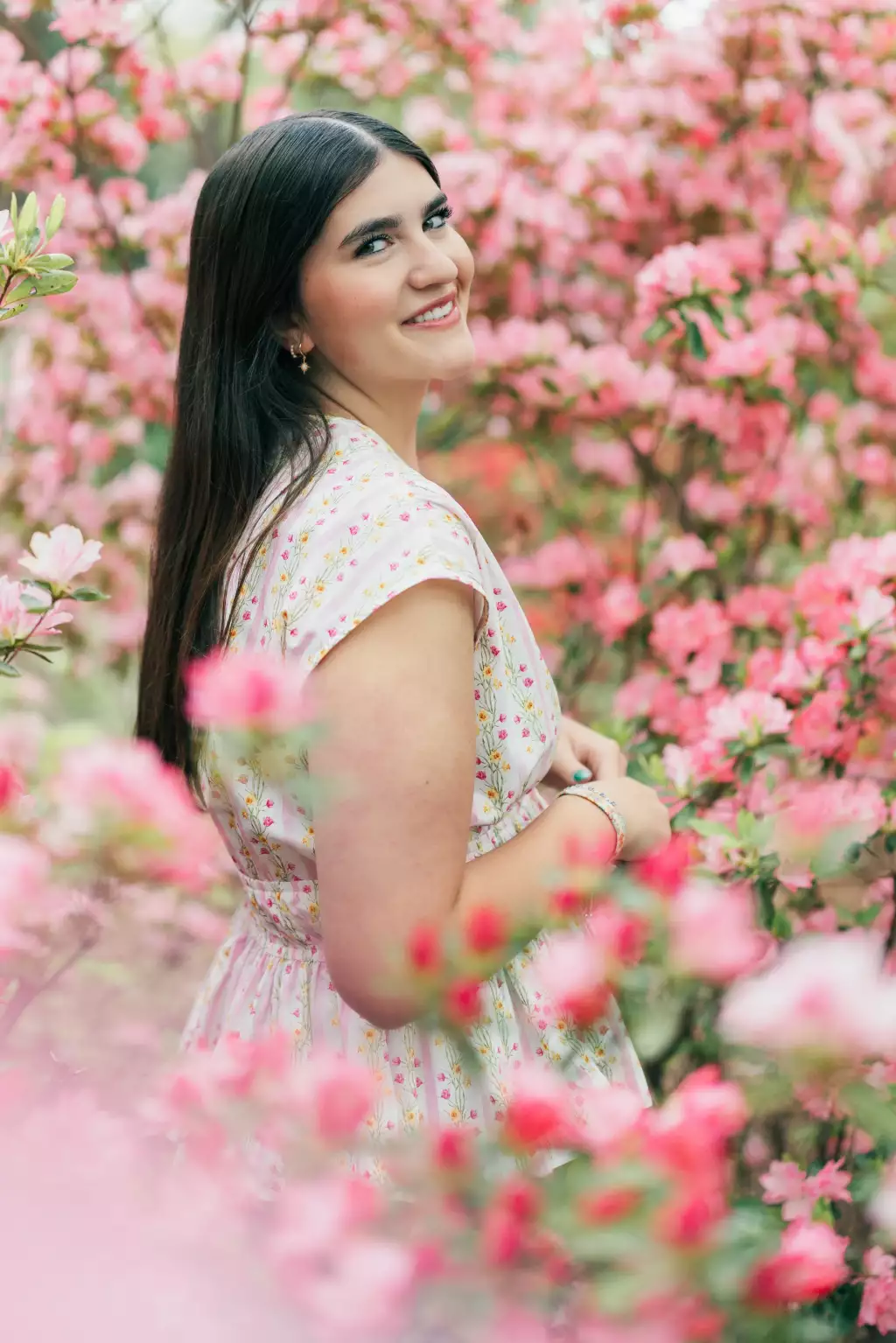 : Woman with long dark hair in a light floral dress standing among bright pink blooming flowers, smiling over her shoulder at the camera.
